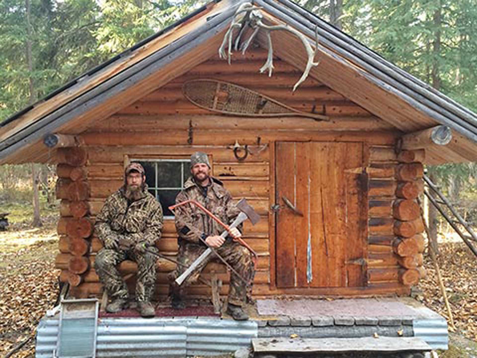 Two men in camouflage sitting in front of a wood cabin 